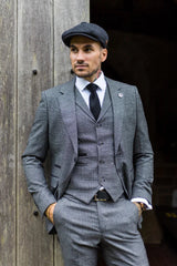 Man in a gray suit and cap standing against a wooden wall.