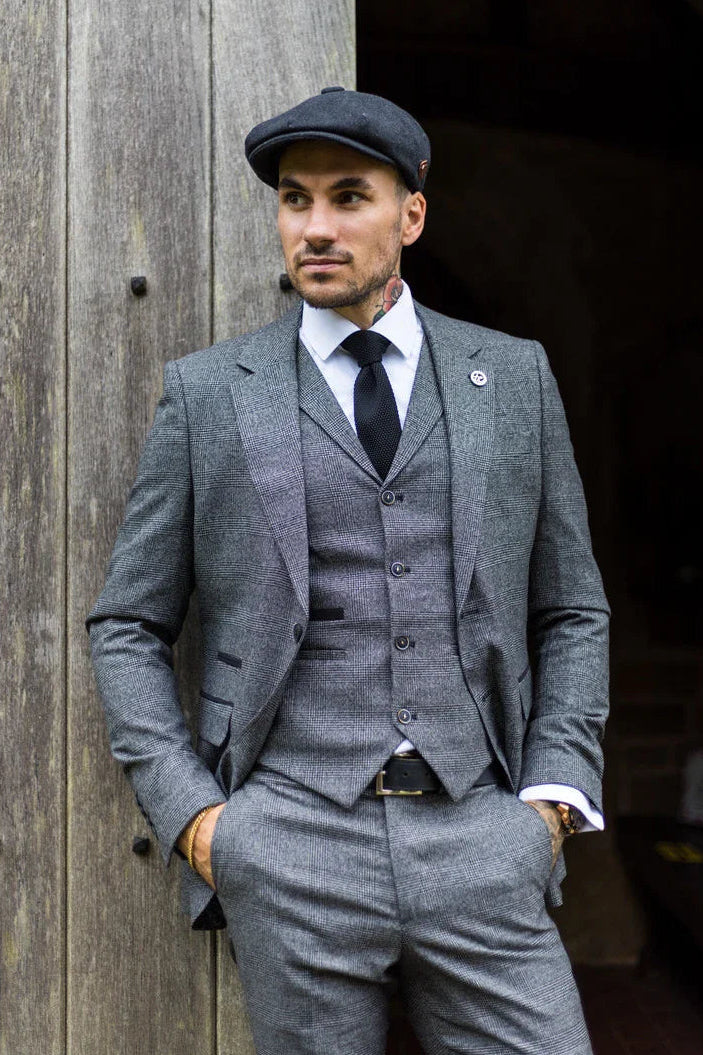 Man in a gray suit and cap standing against a wooden wall.